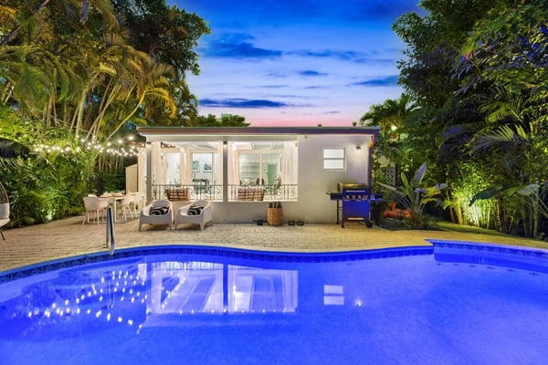 Illuminated swimming pool at dusk with modern house, patio lounge chairs, grill, and tropical landscaping