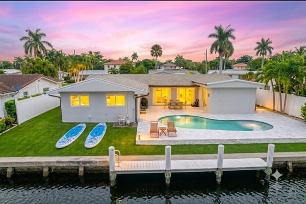 Waterfront home with pool, lit interior, paddleboards on lawn, and dock at sunset with palm trees.